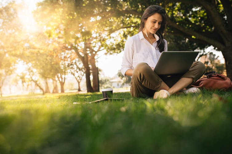 A woman sitting in a park watching something on her laptop, with earphones in.