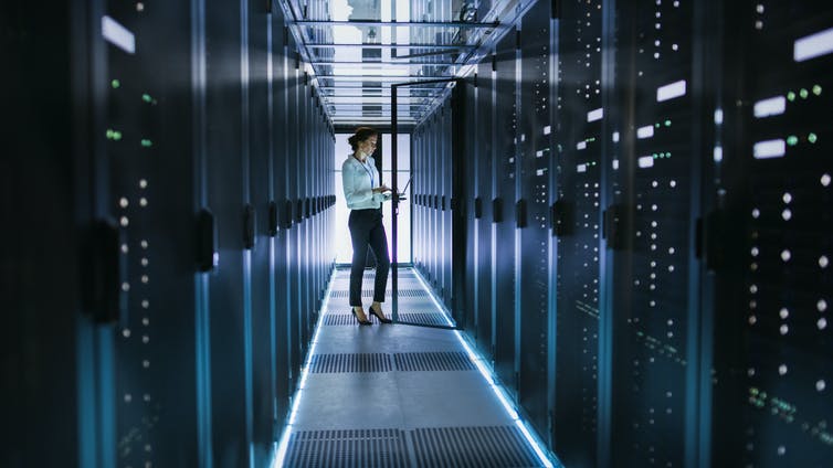 Female Server Technician Stands next to Cabinet in Data Center Corridor with Rows of Rack Servers. She's Running Diagnostics on Her Computer