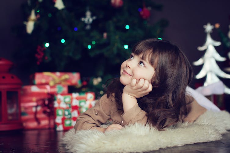 Girl with dark hair lying on the carpet and smiling with Christmas tree in the background.